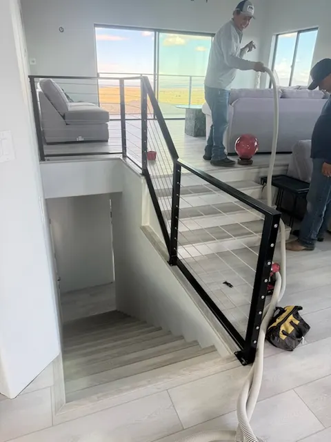 Two installers finishing a black steel cable railing along an indoor stair opening
