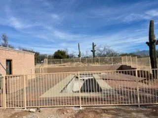 Tan steel picket fence enclosing a desert yard beside a brick building, with saguaros behind