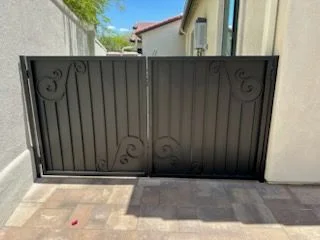 Black steel double gate with vertical bars and scroll accents across a paver drive
