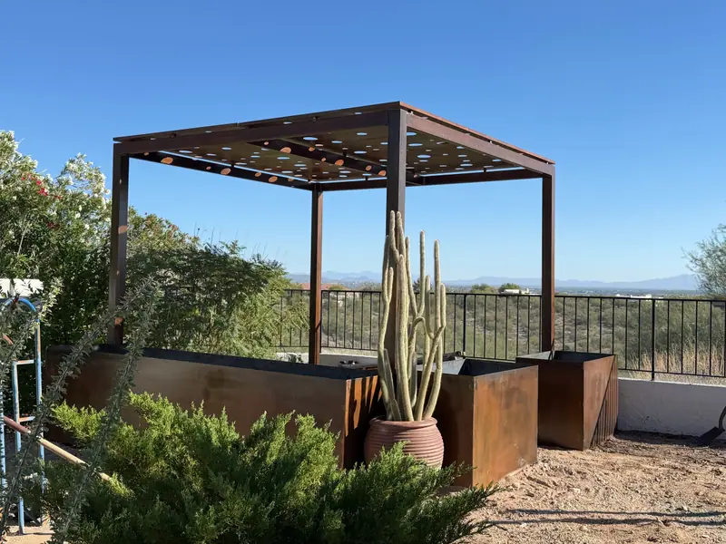 Steel pergola with circle-cut roof panels over rusted steel planter boxes, with the Tucson valley and mountains behind