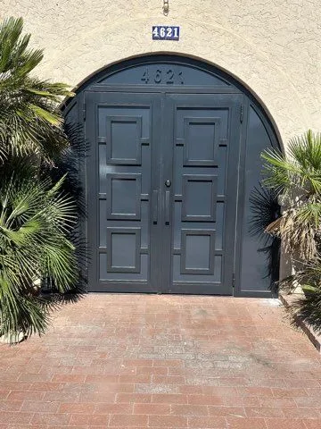 Charcoal steel double doors with raised square panels under a matching arched top, framed by palms over a brick walk