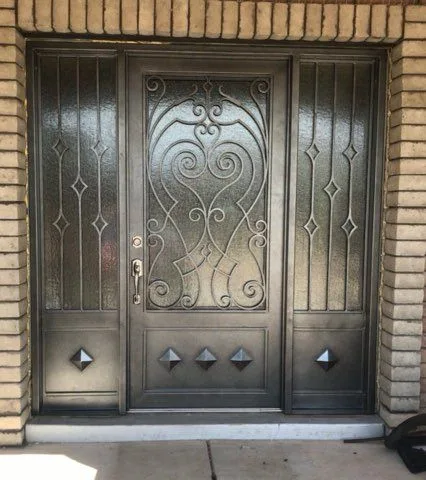 Dark steel door with a scrollwork center, matching sidelights over textured glass and pyramid studs on the lower panels, in a brick entry