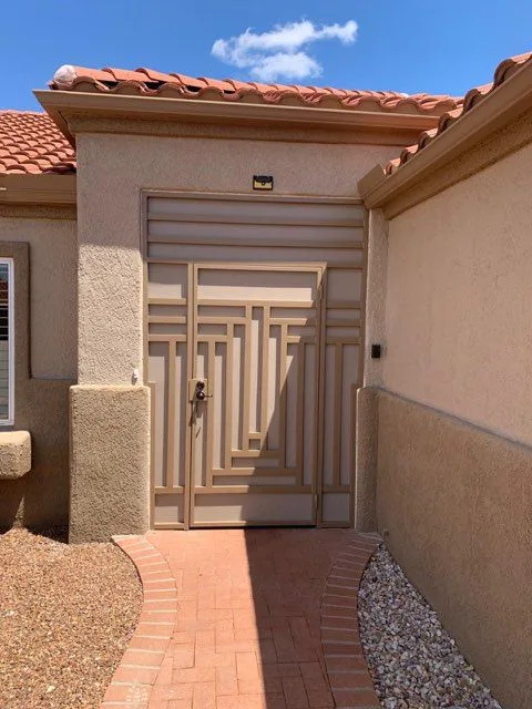 Tan security door with a stepped line pattern and a matching panel above it, at a stucco entry with a brick paver walk