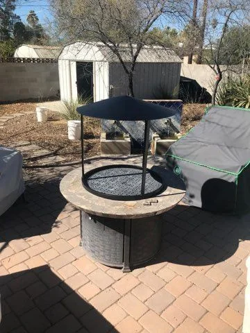 Black steel hood on legs over a round fire table on a brick paver patio