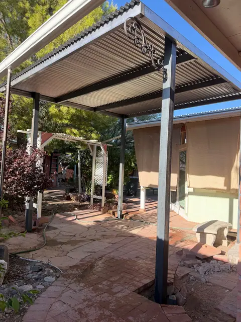 Steel patio cover with a corrugated roof and scroll brackets over a brick courtyard
